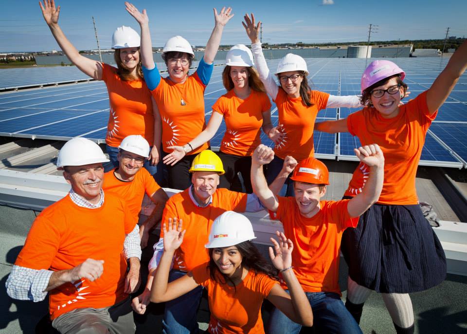 group shot of excited people with their arms in the air on top of a roof with solar panels. people are wearing hard hats and bright orange shirts