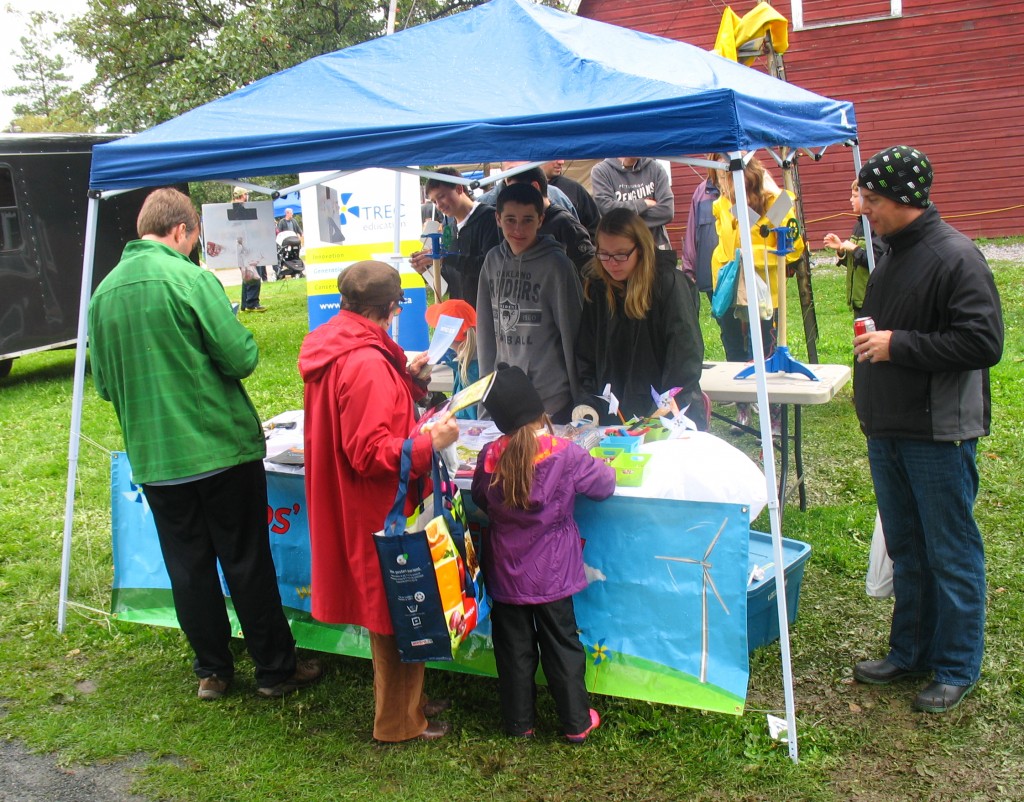 Volunteers stand underneath a blue pop-up tent, and answer questions about renewable energy and energy cosnervation as a young girl plays with games. Three adults are reading information from the table.
