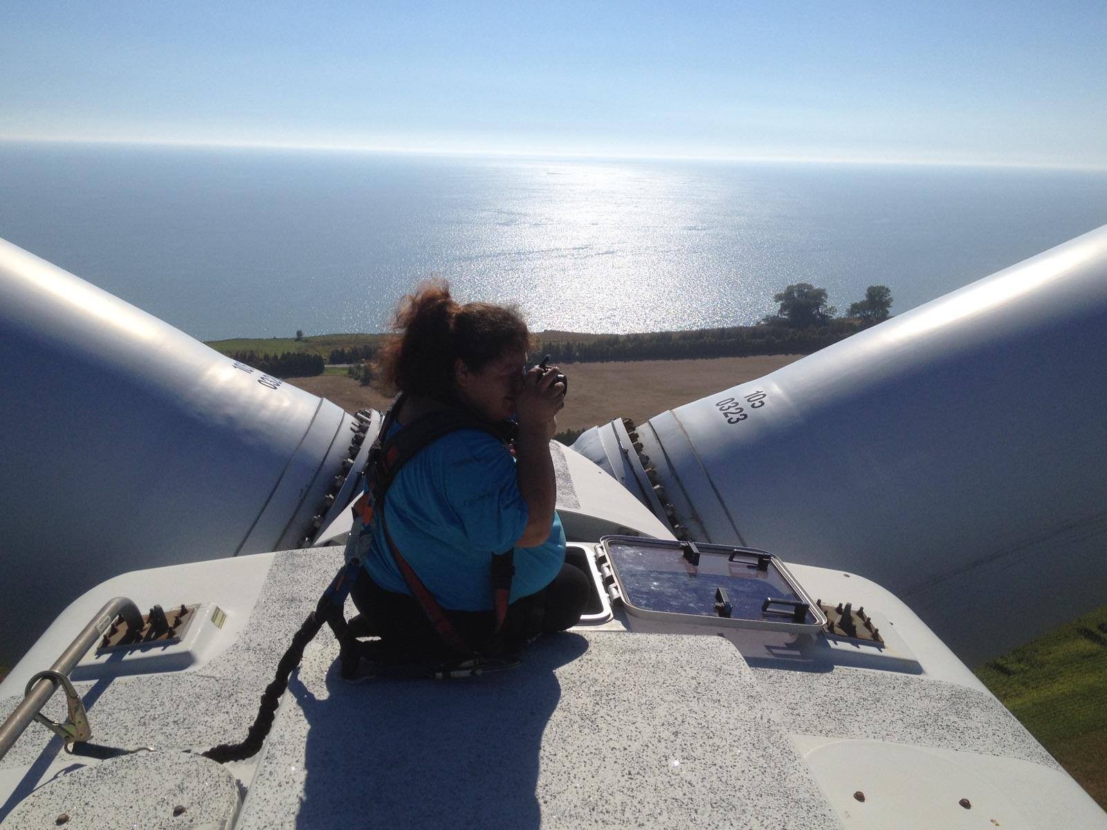 A women in a safety harness takes a photo while crouching outside on the top of a wind turbine, with a view of the lake.