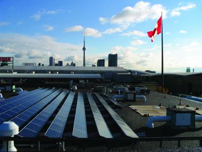 photo of the solar array on top of the Horse Palace roof in Toronto. CN Tower is in the background with the city skyline and a large Canadian Flag waves in the wind on the right.