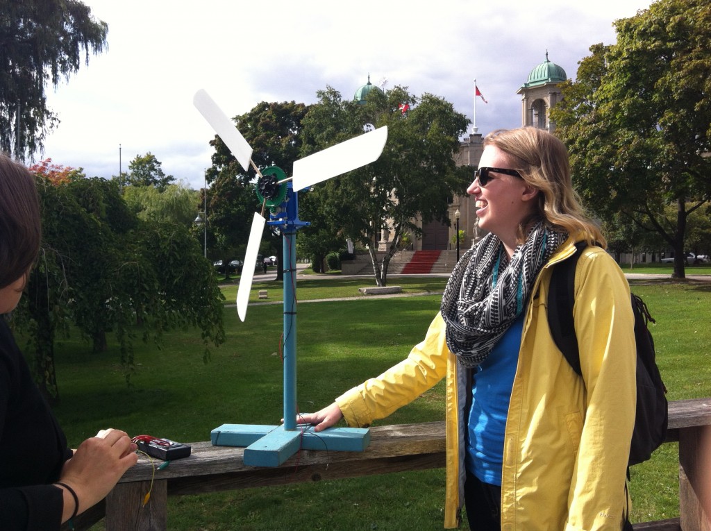 Woman wearing sunglasses smiles while standing next to a model wind turbine outside on a sunny day
